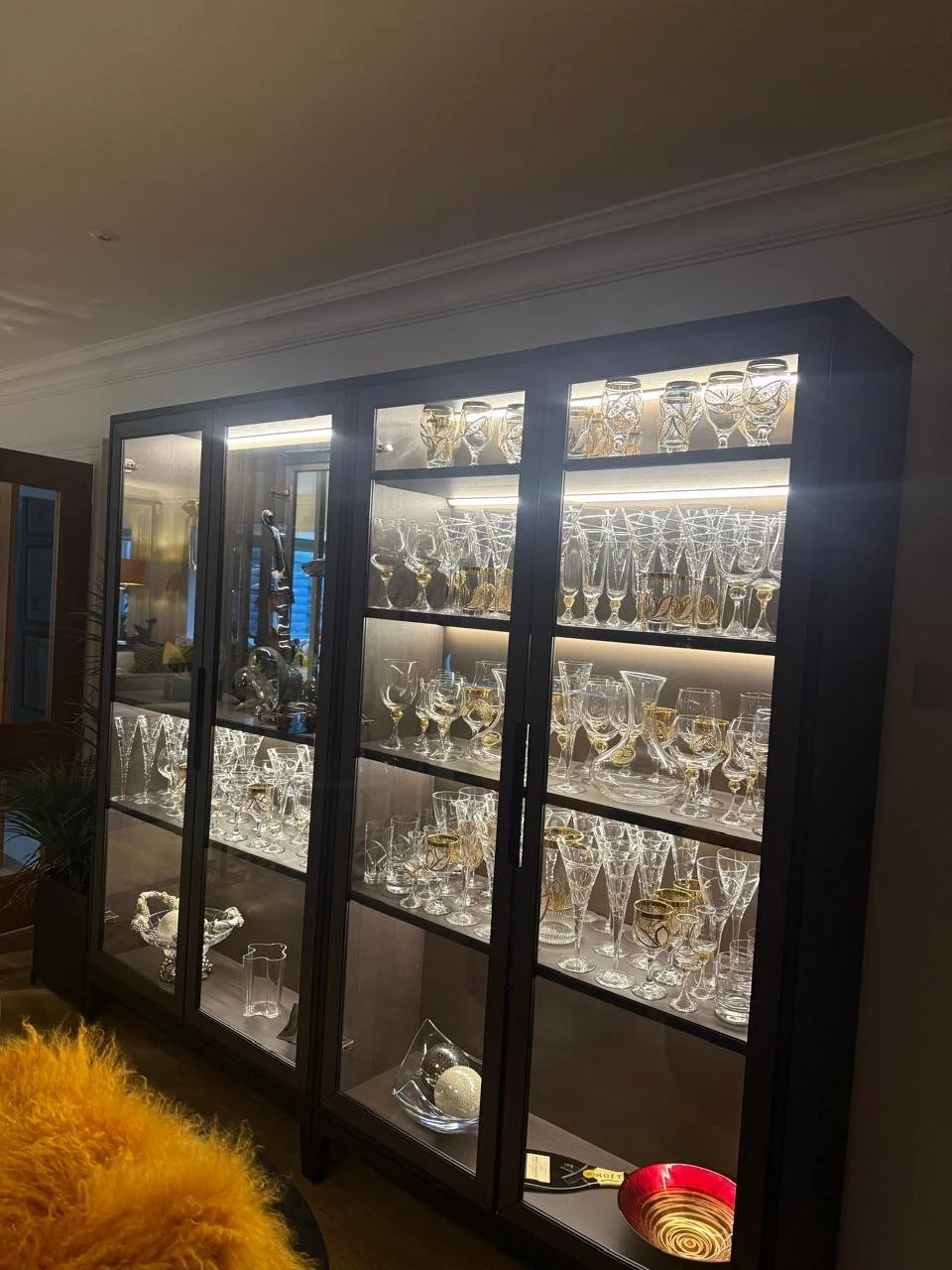 Display cabinet filled with glassware including glasses, bowls, and decorative pieces, with internal lighting highlighting the items.