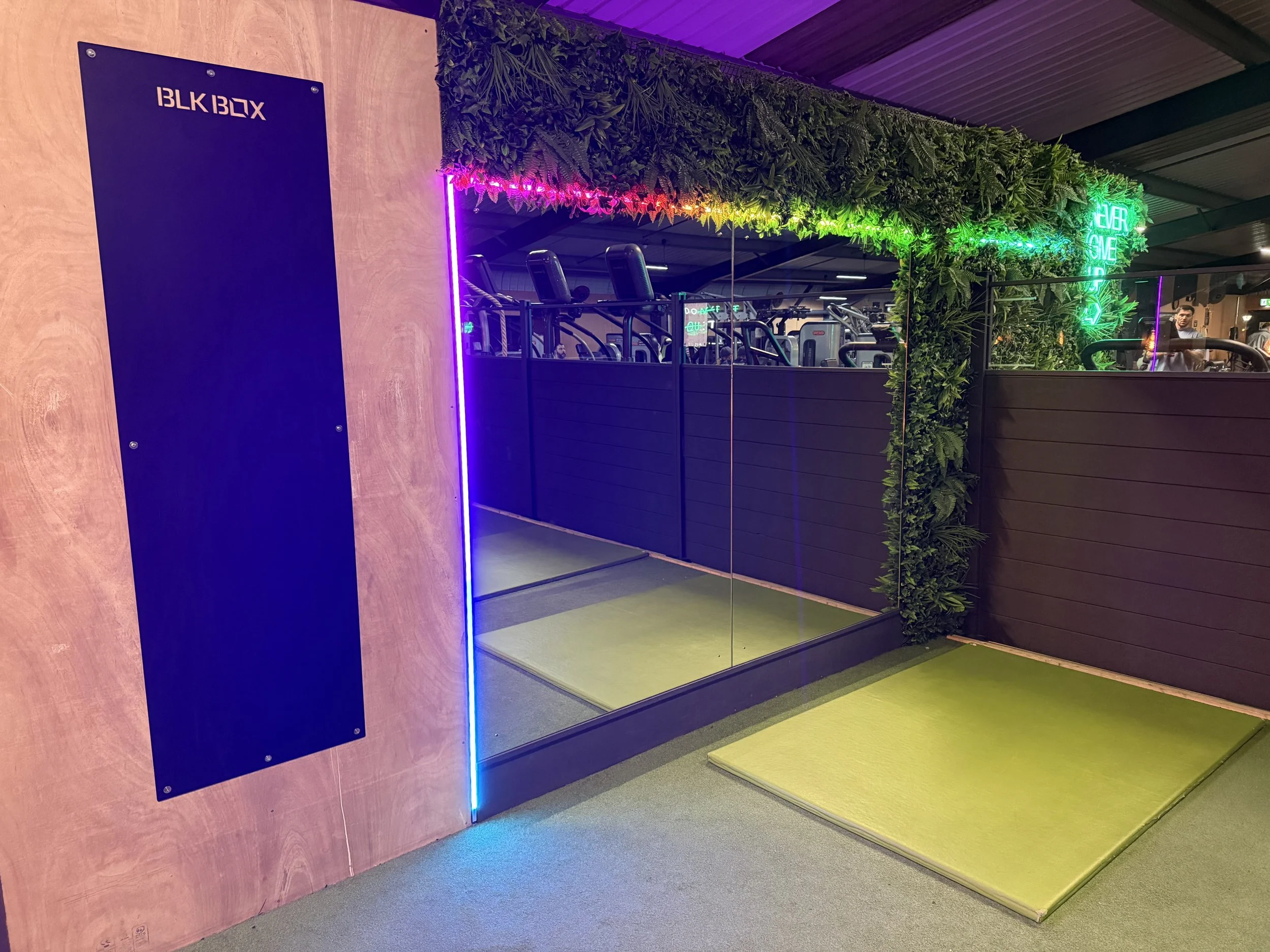 Indoor golf simulation bay with a mirror wall, green turf mat, and contemporary decor including vertical greenery, neon signage, and bright lighting.