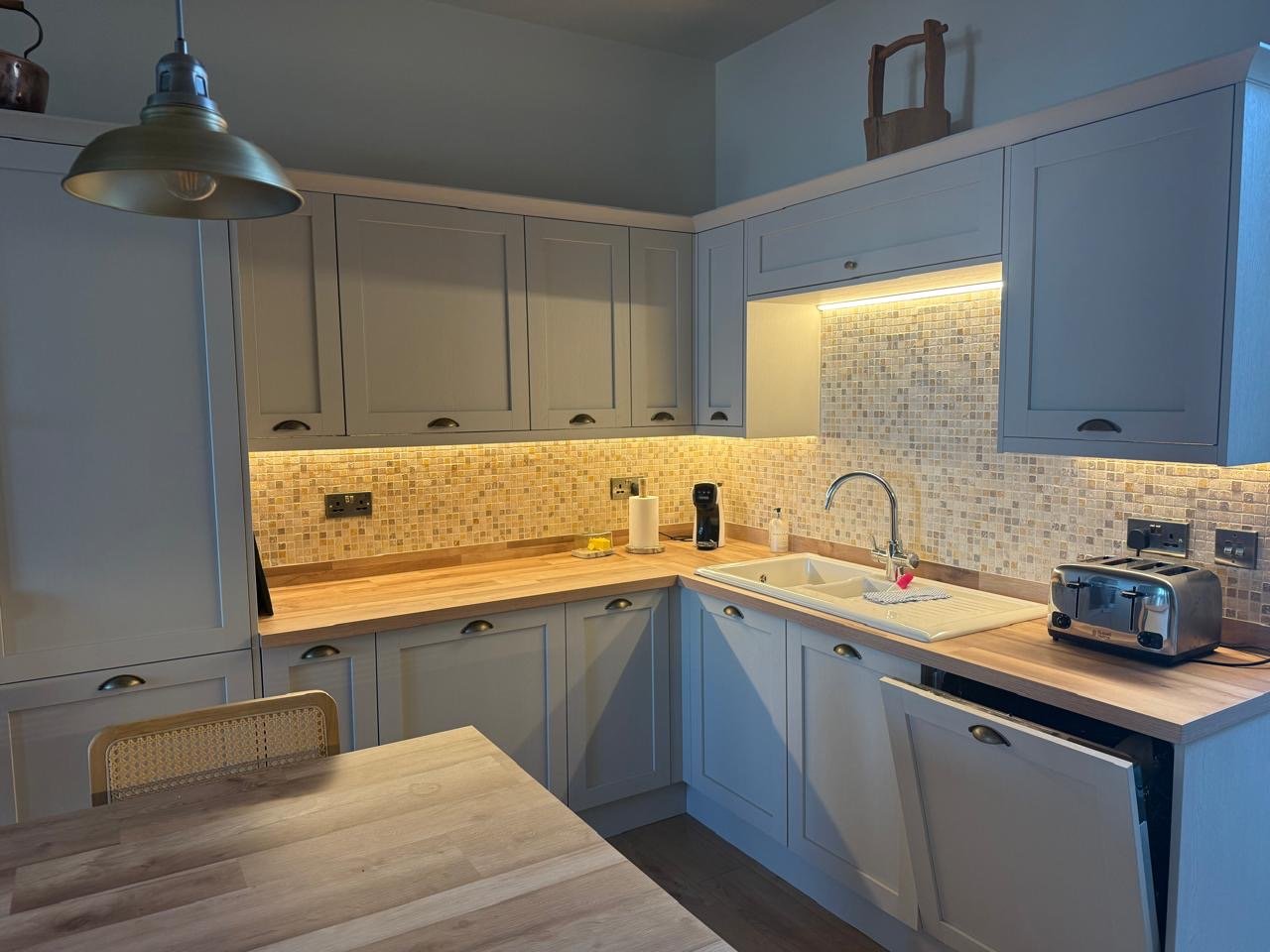 Kitchen with beige cabinets, wooden countertop, mosaic tile backsplash, stainless steel sink, under-cabinet lighting, small kitchen appliances, and wooden flooring.