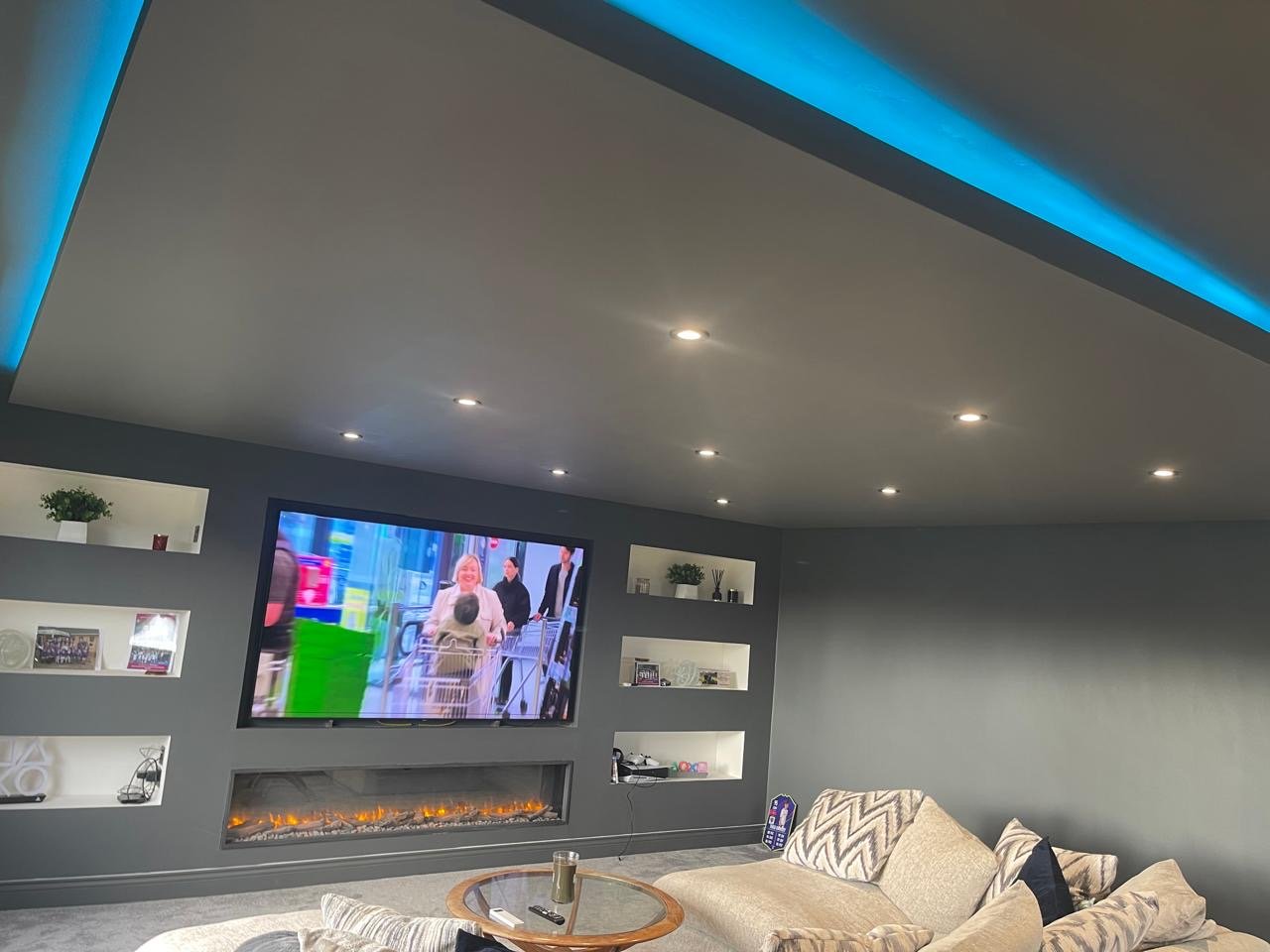 Living room with gray walls and built-in shelves, a flat-screen TV displaying a scene with people at an airport, an electric fireplace below the TV, a beige sectional sofa with patterned pillows, and a round wooden coffee table with a can and remote on it. Recessed ceiling lights and blue accent lighting on the ceiling.