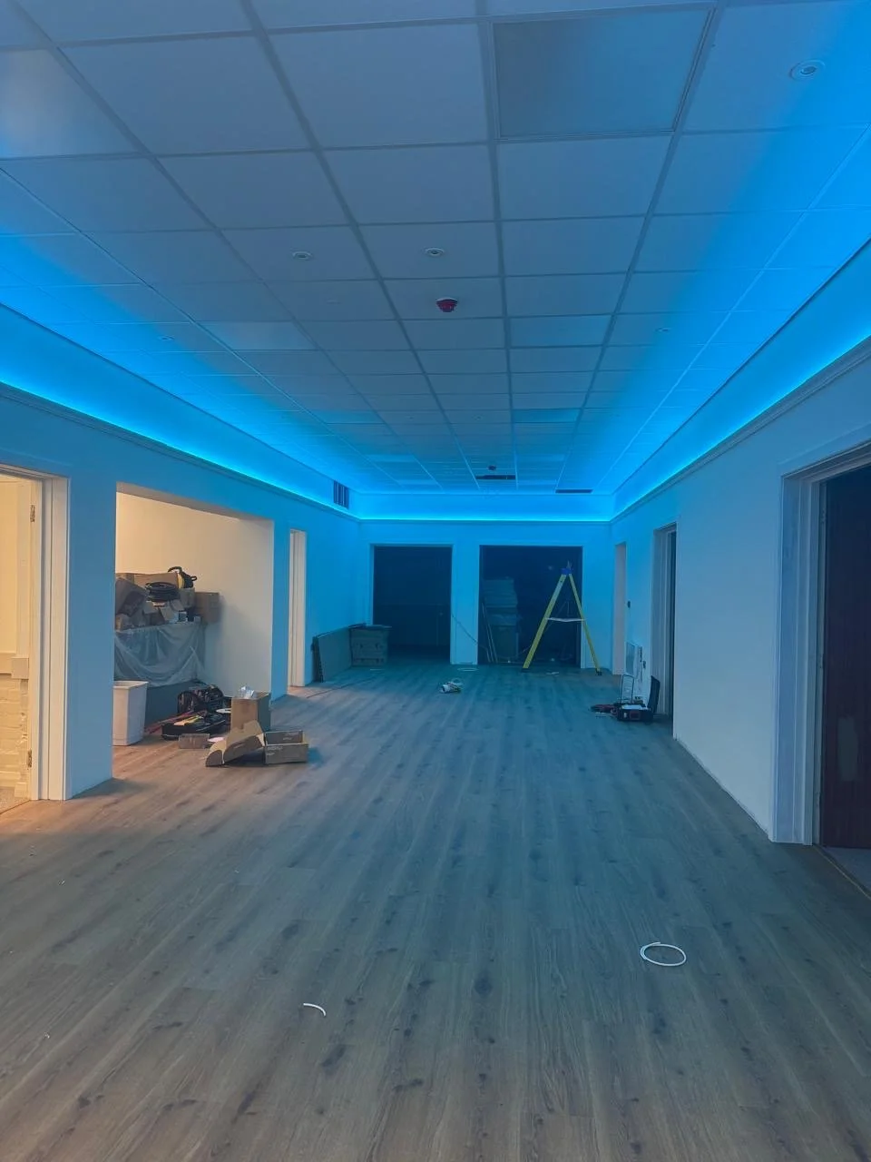 Empty room with wooden flooring, illuminated by blue LED lighting along the ceiling edges, under construction with tools and supplies scattered around.