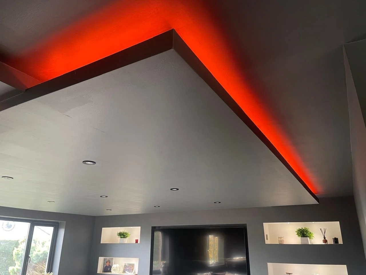 Ceiling with recessed lights and a strip of red LED lighting on the edge, above a modern living room with shelves and a television.