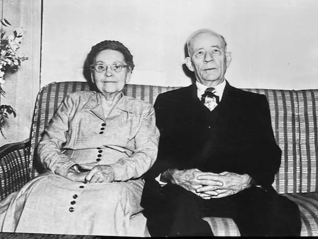 An elderly woman and man sitting on a striped sofa in a black-and-white photo.