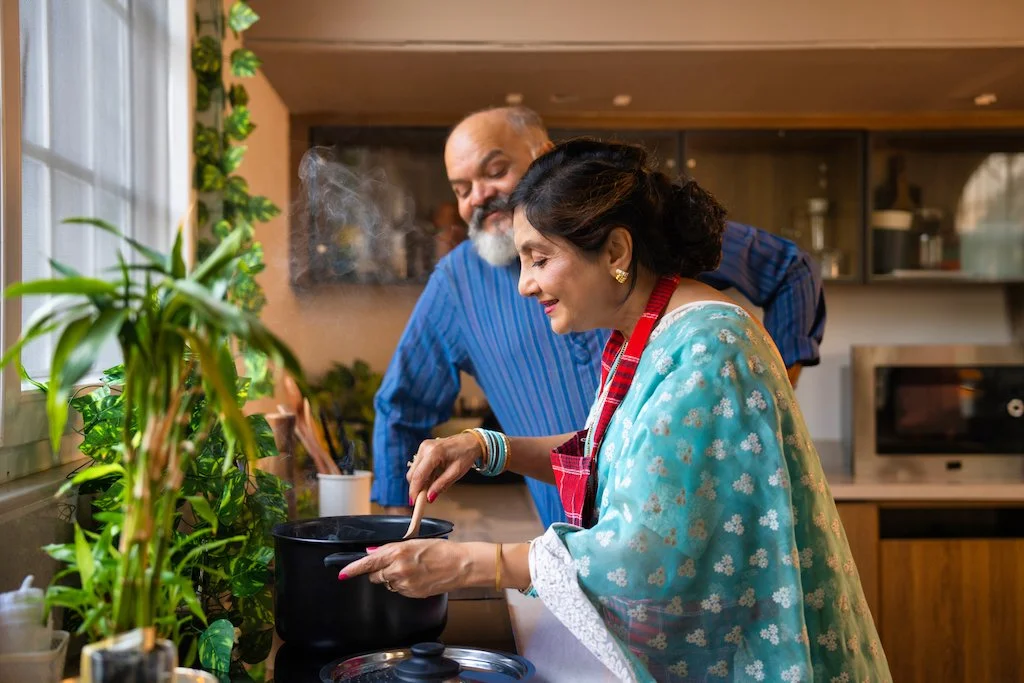 A woman and a man cooking together in a kitchen, with the woman stirring a pot on the stove and smiling, and plants on the windowsill.