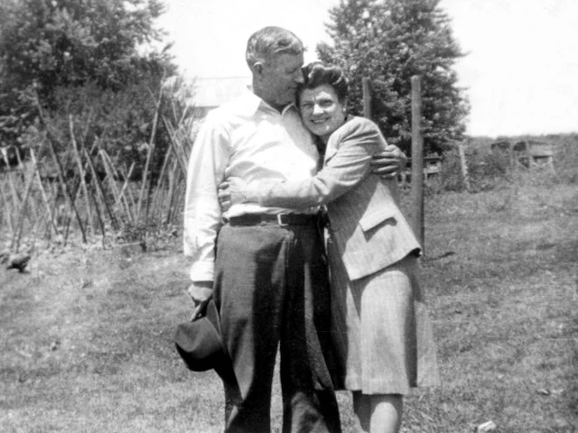 A man and woman affectionately embracing in a grassy outdoor setting, with trees and a garden structure in the background. The man is holding a hat.