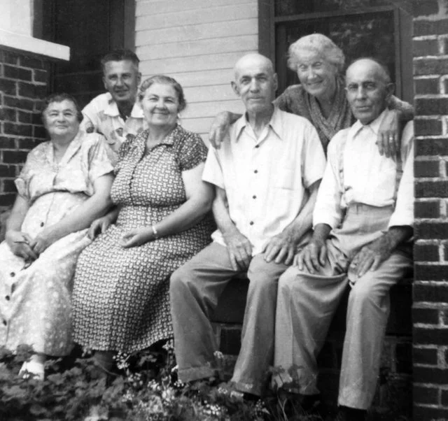 A black and white photo of six elderly people sitting together on a porch, with brick and siding on the building in the background.