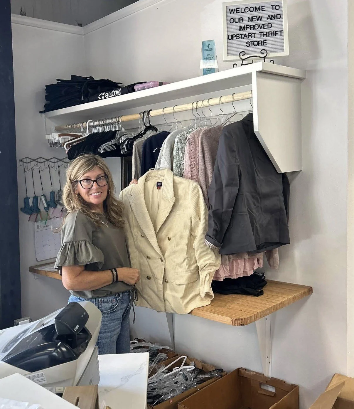 A volunteer hangs clothing beneath a sign welcoming shoppers to the new and improved UpStart Thrift Store in Farmington, Arkansas
