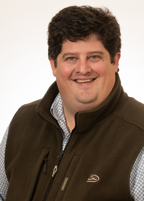 Portrait of a smiling man with dark curly hair wearing a brown vest over a blue checkered shirt, against a plain light background.
