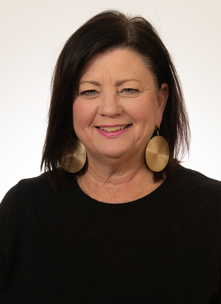 A middle-aged woman with dark brown hair, wearing gold earrings and a black top, smiling against a light background.