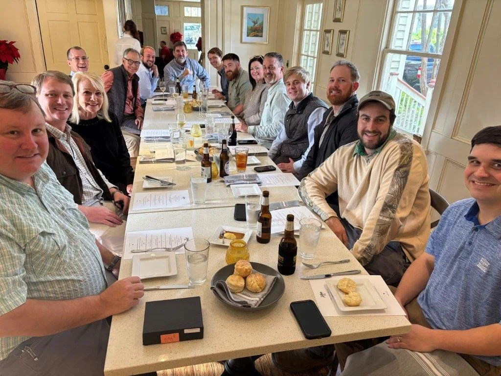 Group of people sitting around a long dining table in a well-lit room, enjoying food and drinks during a gathering.