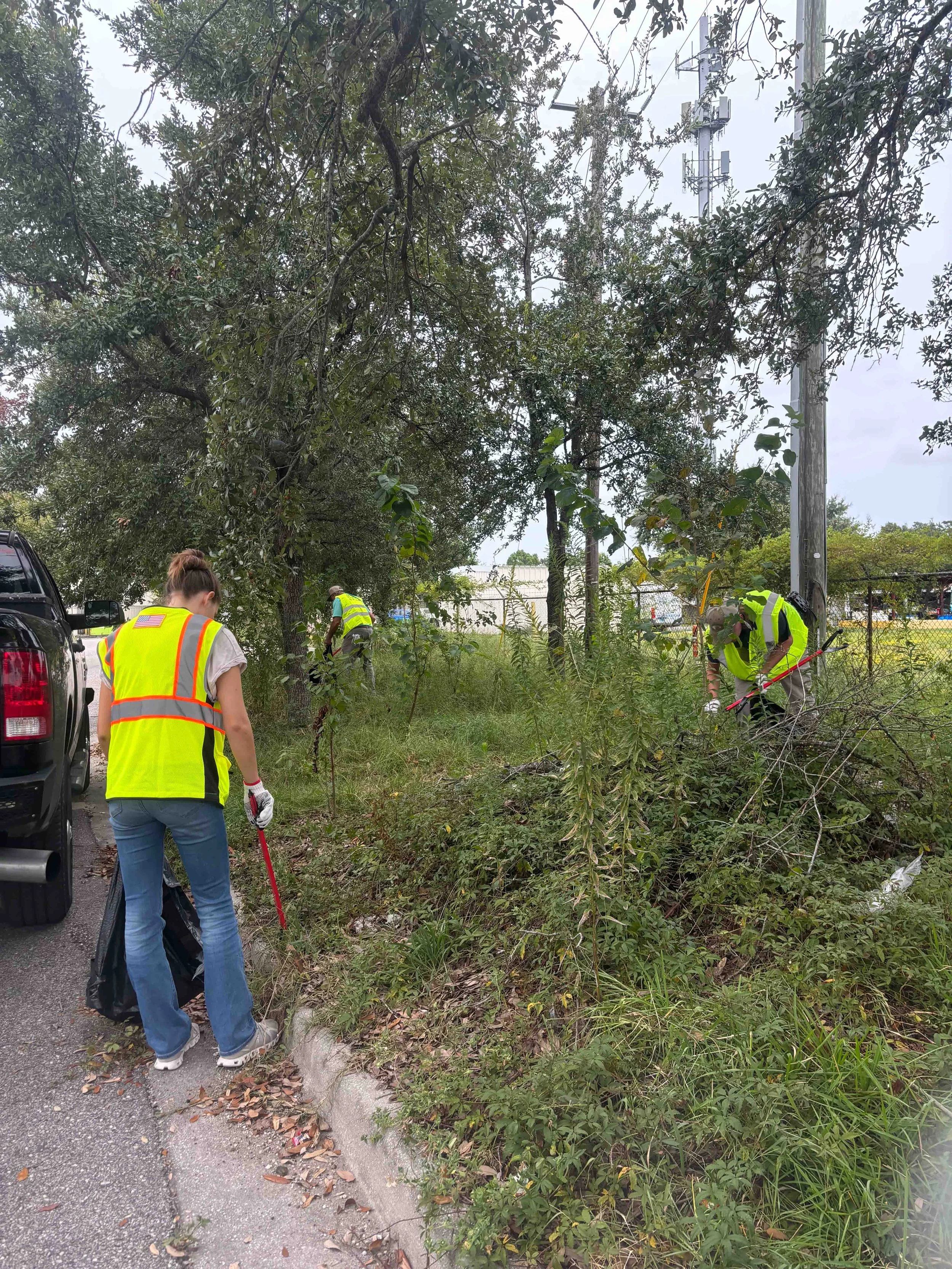 Three people in yellow safety vests are working outdoors near a utility pole, trimming trees and bushes along a grassy area next to a street, with a truck parked nearby.
