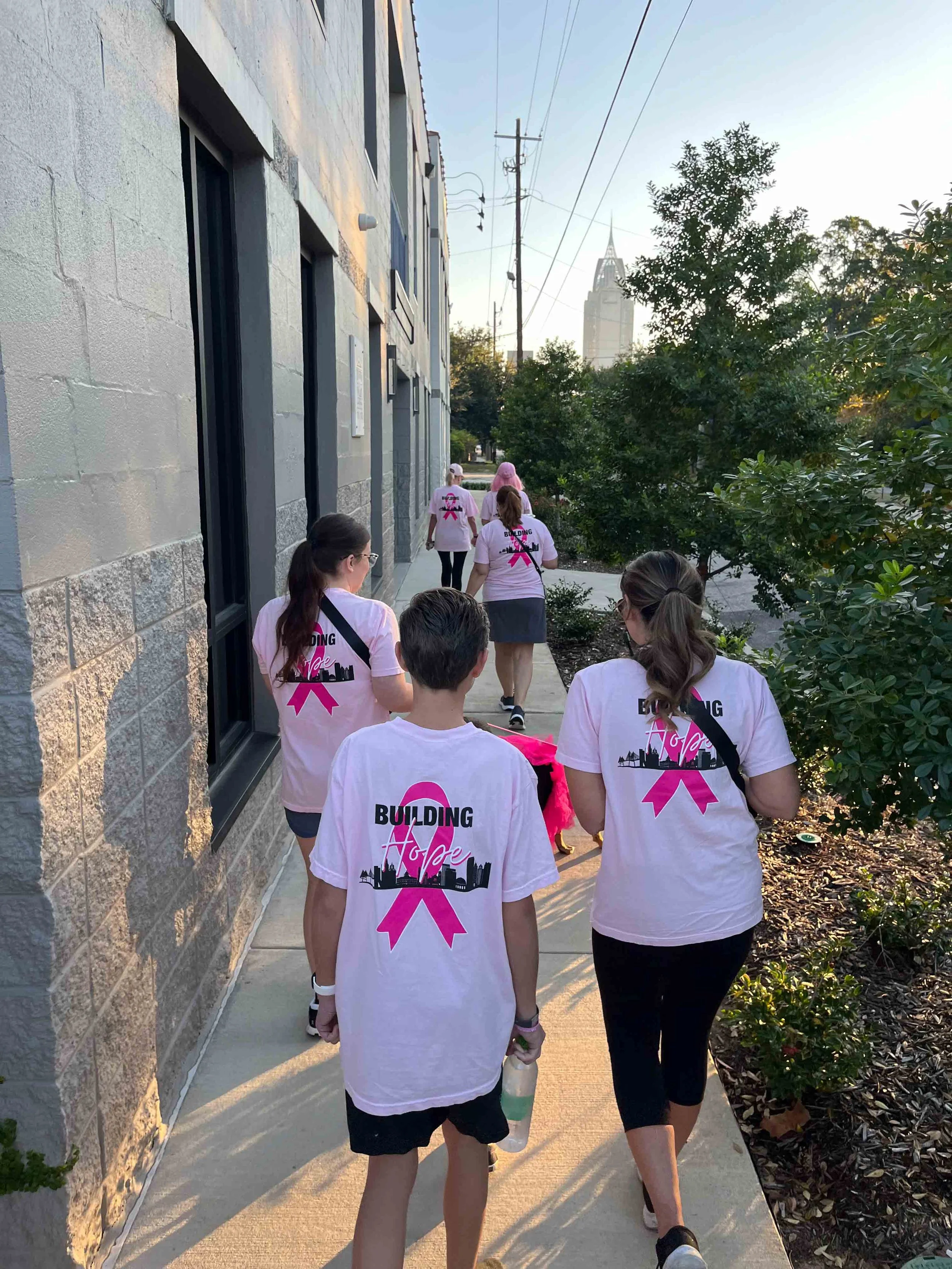 Group of people wearing pink ribbons and matching t-shirts participating in a walk or march along a sidewalk in an urban area, with a tall building visible in the distance and trees on the right side.