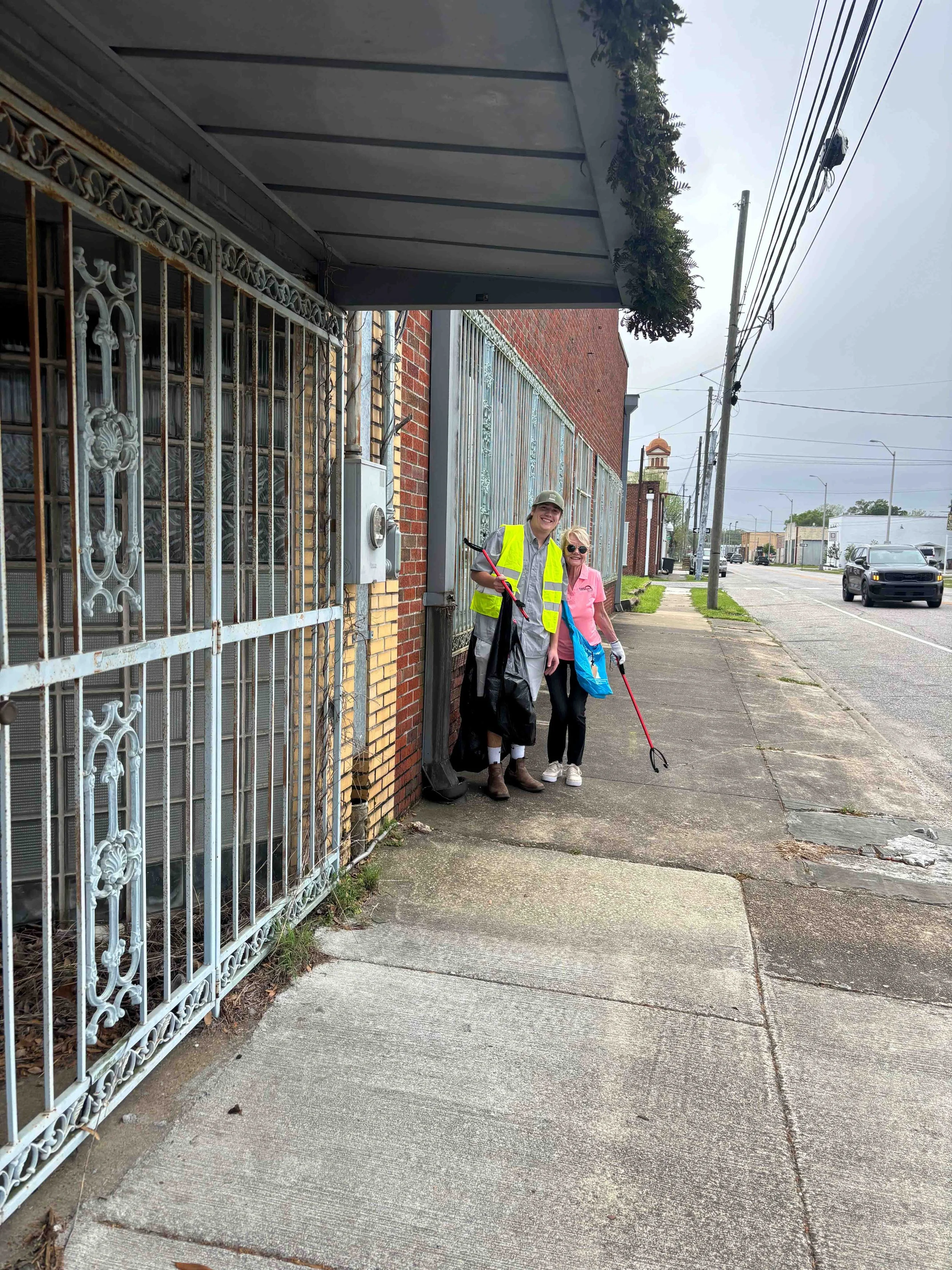 Two women standing on a sidewalk, one wearing a high-visibility vest and holding trash pick-up tools, smiling at the camera.