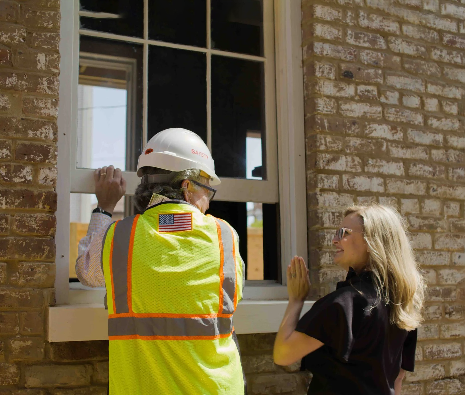 A construction worker wearing a high visibility vest and a white safety helmet talking to a woman near a brick wall. The worker has an American flag patch on the back of the vest and is holding a window frame.