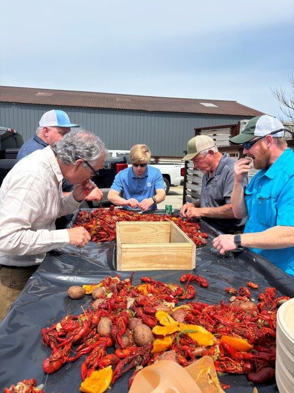 Six people gathered around a table covered with crawfish, potatoes, and yellow bell peppers, outdoors on a cloudy day, with industrial buildings in the background.