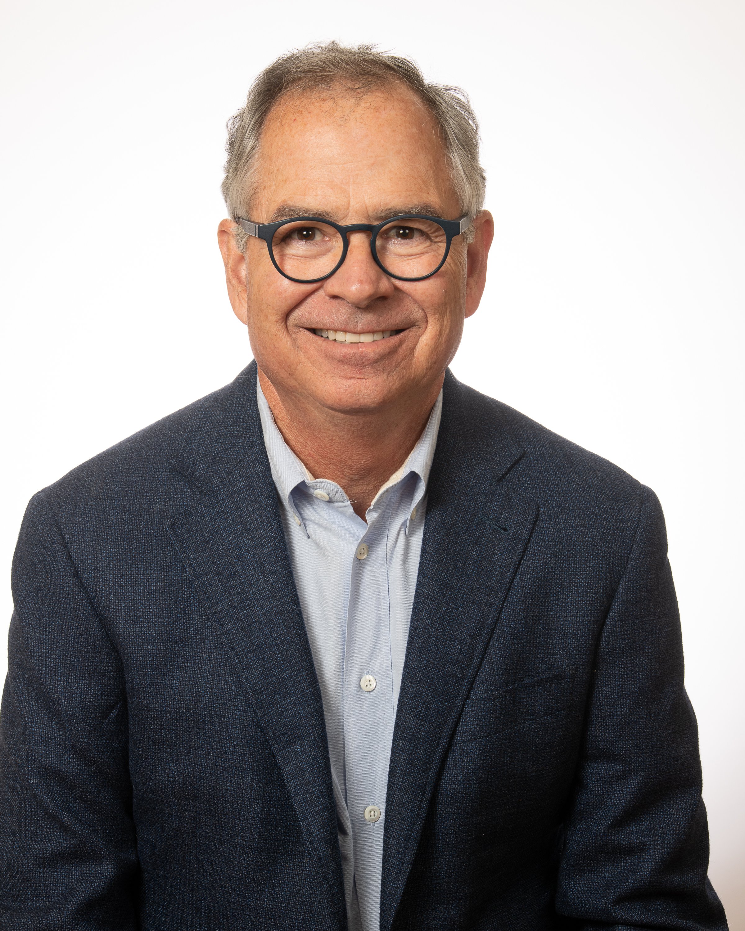 Headshot of a middle-aged man with gray hair, glasses, and a friendly smile, wearing a light-colored button-up shirt.