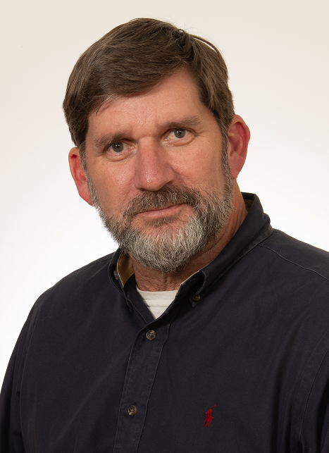 A middle-aged man with brown hair and a beard, wearing a dark button-down shirt with a small red logo, posing against a plain white background.