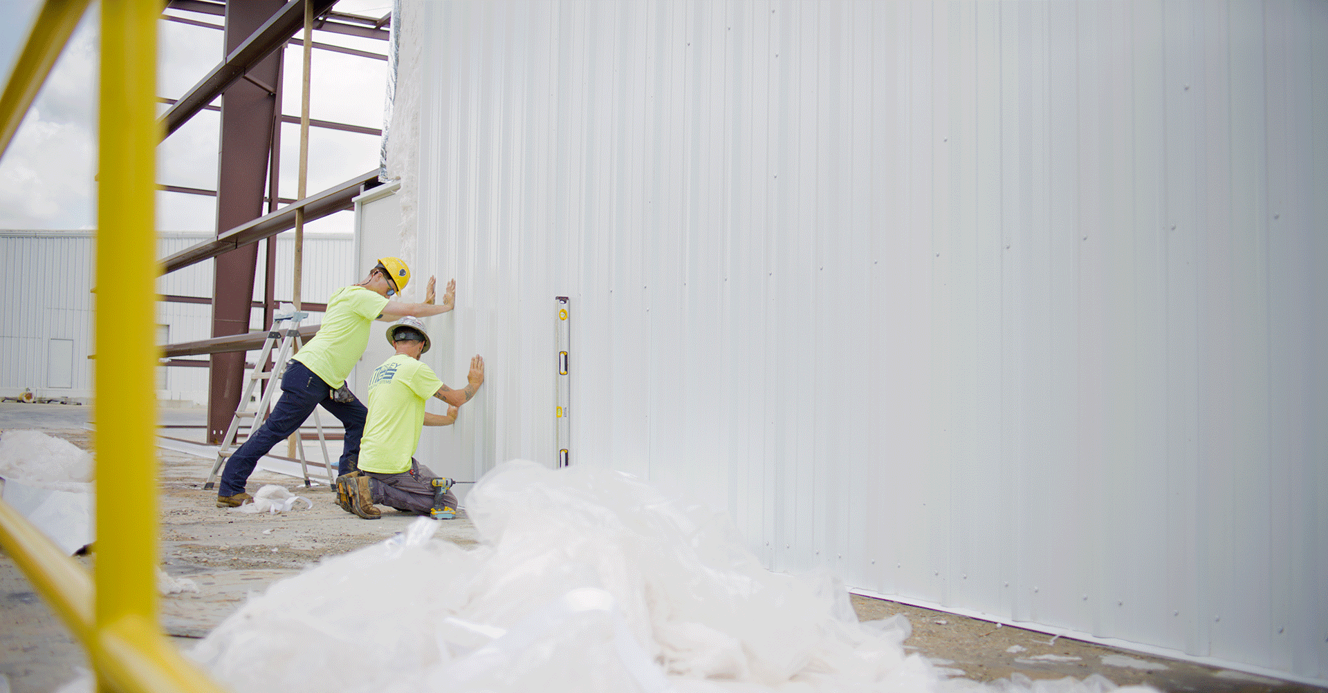Two construction workers, both wearing yellow safety helmets and high-visibility shirts, are installing or inspecting a large white metal wall. One worker is standing on a ladder, pressing against the wall, while the other is kneeling and using a level. Construction materials and tools are on the ground, and part of a yellow scaffolding is visible in the foreground.