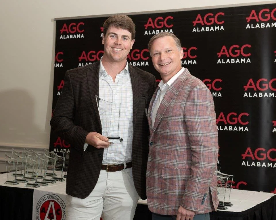 Two men standing together smiling in front of a black backdrop with red and white AGC Alabama logos, one holding a small award or plaque.