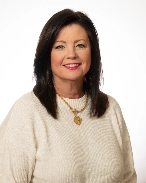 A middle-aged woman with dark brown hair, wearing gold earrings and a black top, smiling against a light background.