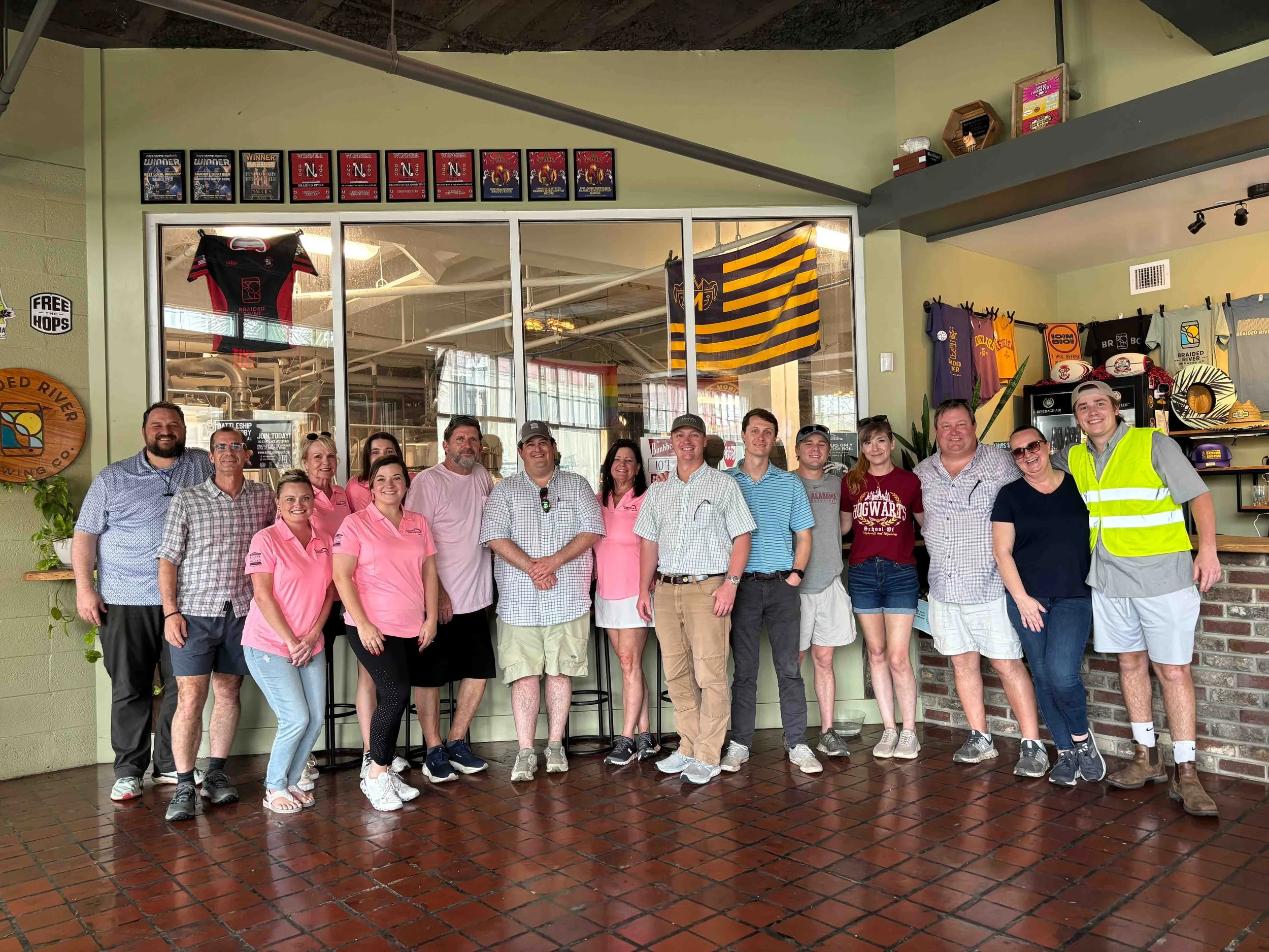 Group of people standing together indoors, some wearing pink shirts, in front of a large window with decorations and awards on the wall.