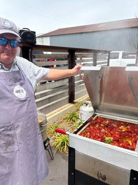 A man wearing a gray apron and cap stands next to a large stainless steel outdoor cooker filled with cooking crawfish. The man is holding a utensil, and in the background there are plants, a trash can, and a wooden fence.