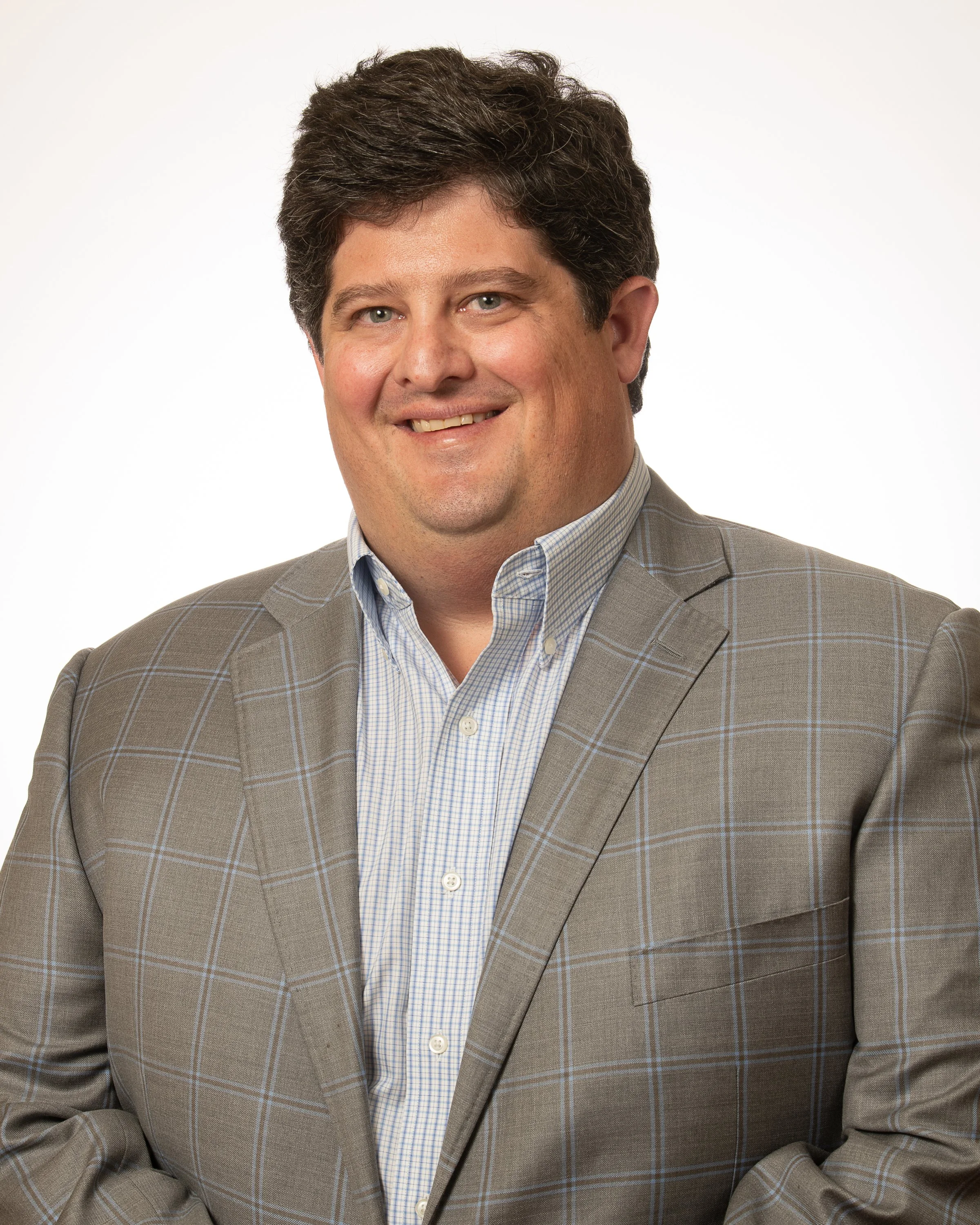 Portrait of a smiling man with dark curly hair wearing a brown vest over a blue checkered shirt, against a plain light background.