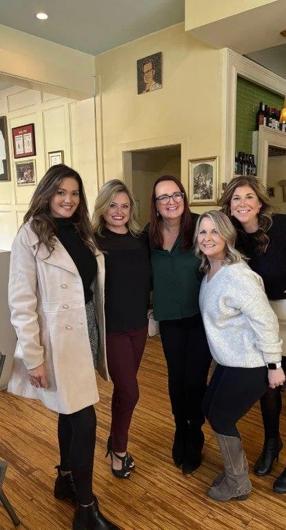 Five women standing together in a cozy room, smiling for the photo.