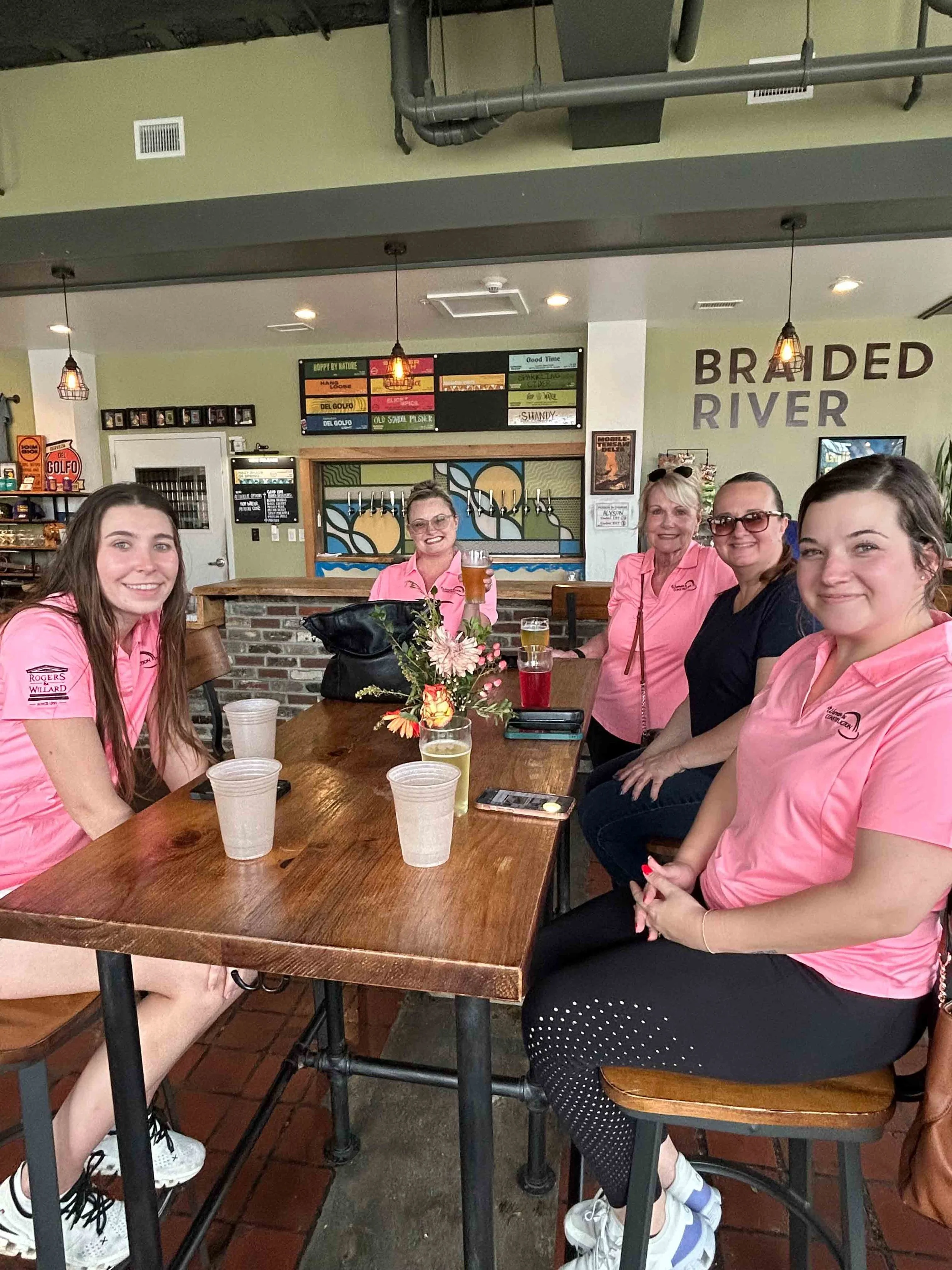 Group of five women sitting at a table in a restaurant, smiling, with drinks in front of them, some wearing pink shirts.