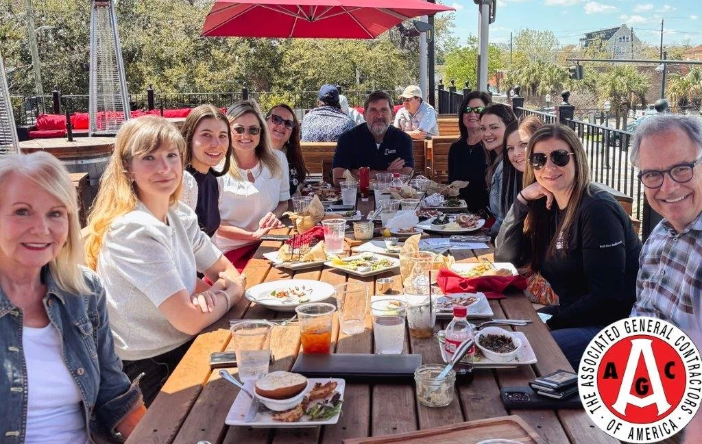 People sitting at a long outdoor wooden table having a meal on a sunny day, with some pizza, drinks, and condiments.