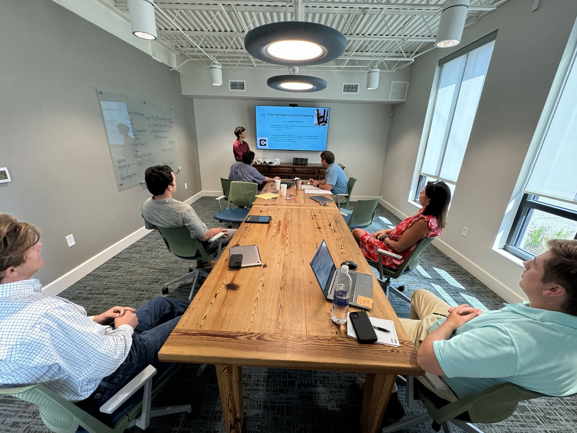 A group of people participating in a business meeting or presentation in a conference room with large windows, a projector screen, and a wooden table.