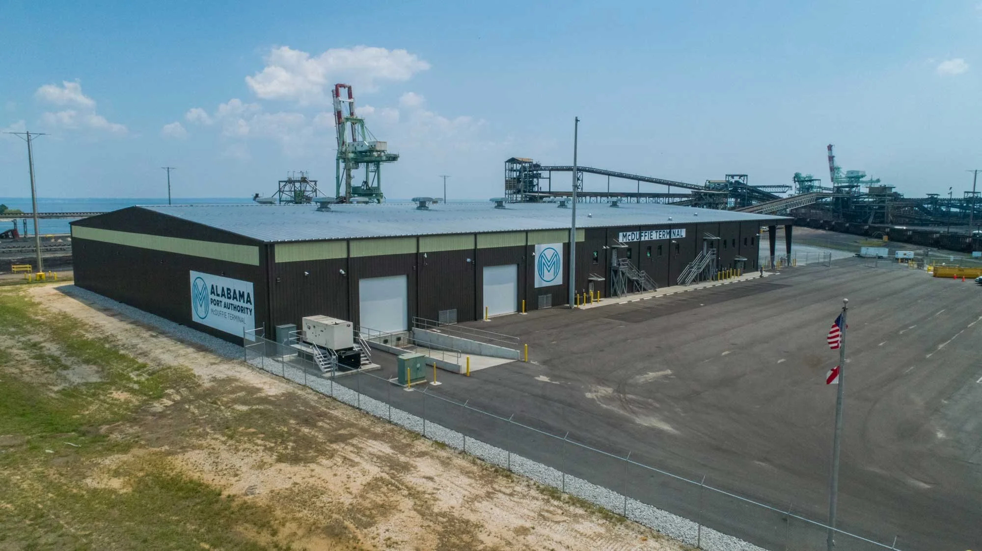 A large industrial building with a sign that says Alabama Port Authority McDuffie Terminal, surrounded by an empty parking lot, with industrial cranes and equipment in the background.