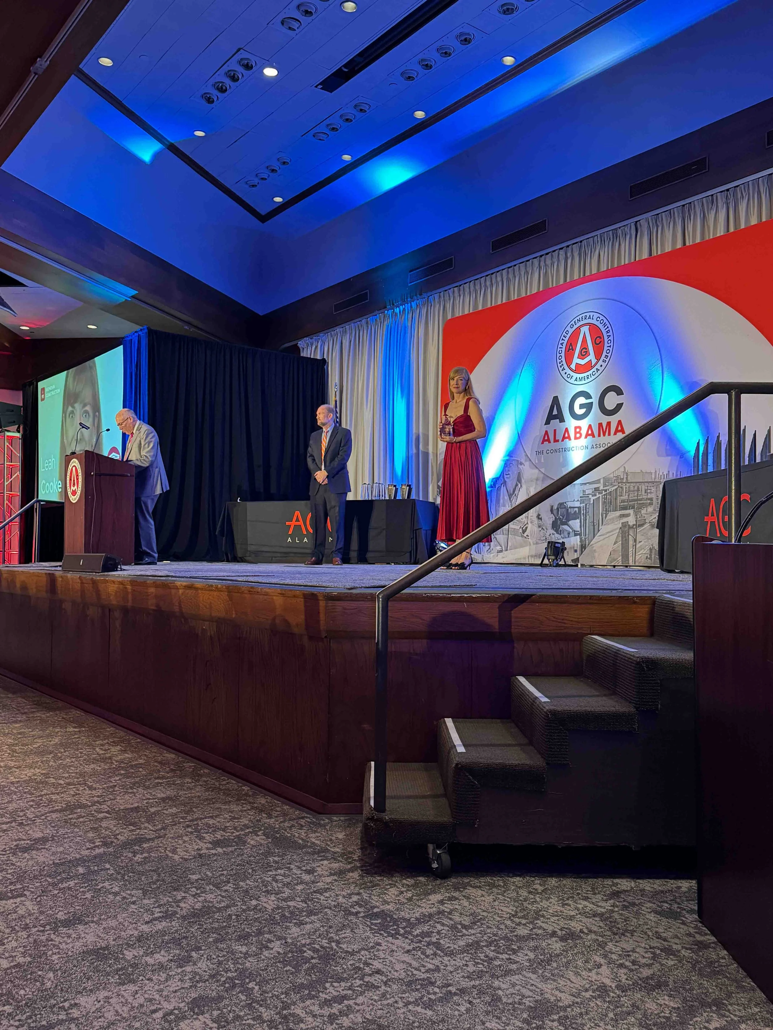 A stage at an Alabama Construction Association event with four people standing, including a woman in a red dress, and a large screen and backdrop displaying the AGC Alabama logo.