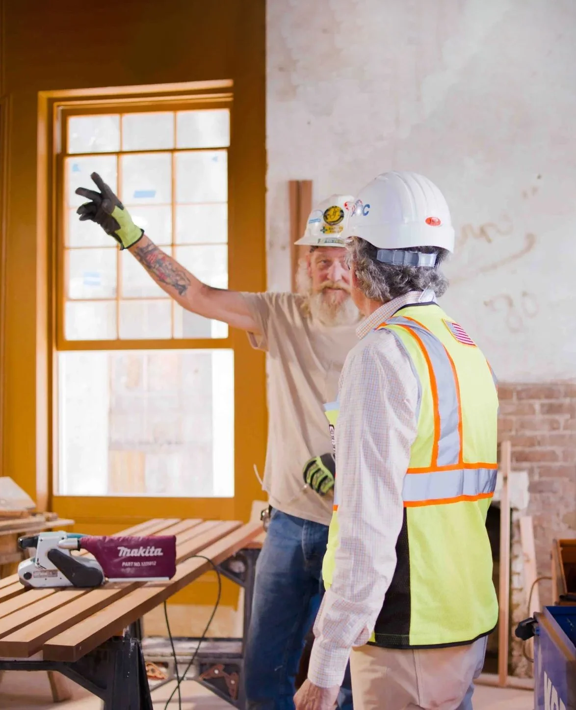 Two construction workers wearing safety helmets and vests are inside a building, with one pointing toward the window while the other listens.