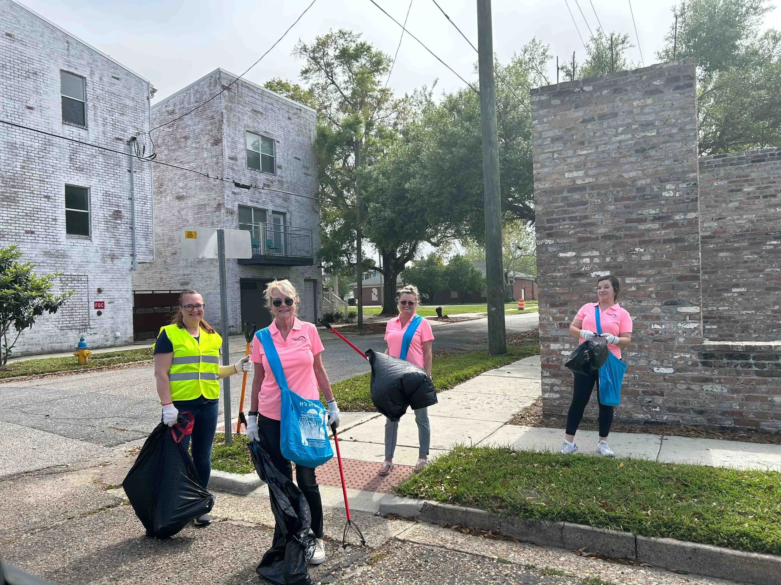 Four women participating in a community cleanup, wearing gloves and holding trash bags, standing on a sidewalk next to a brick wall and trees.