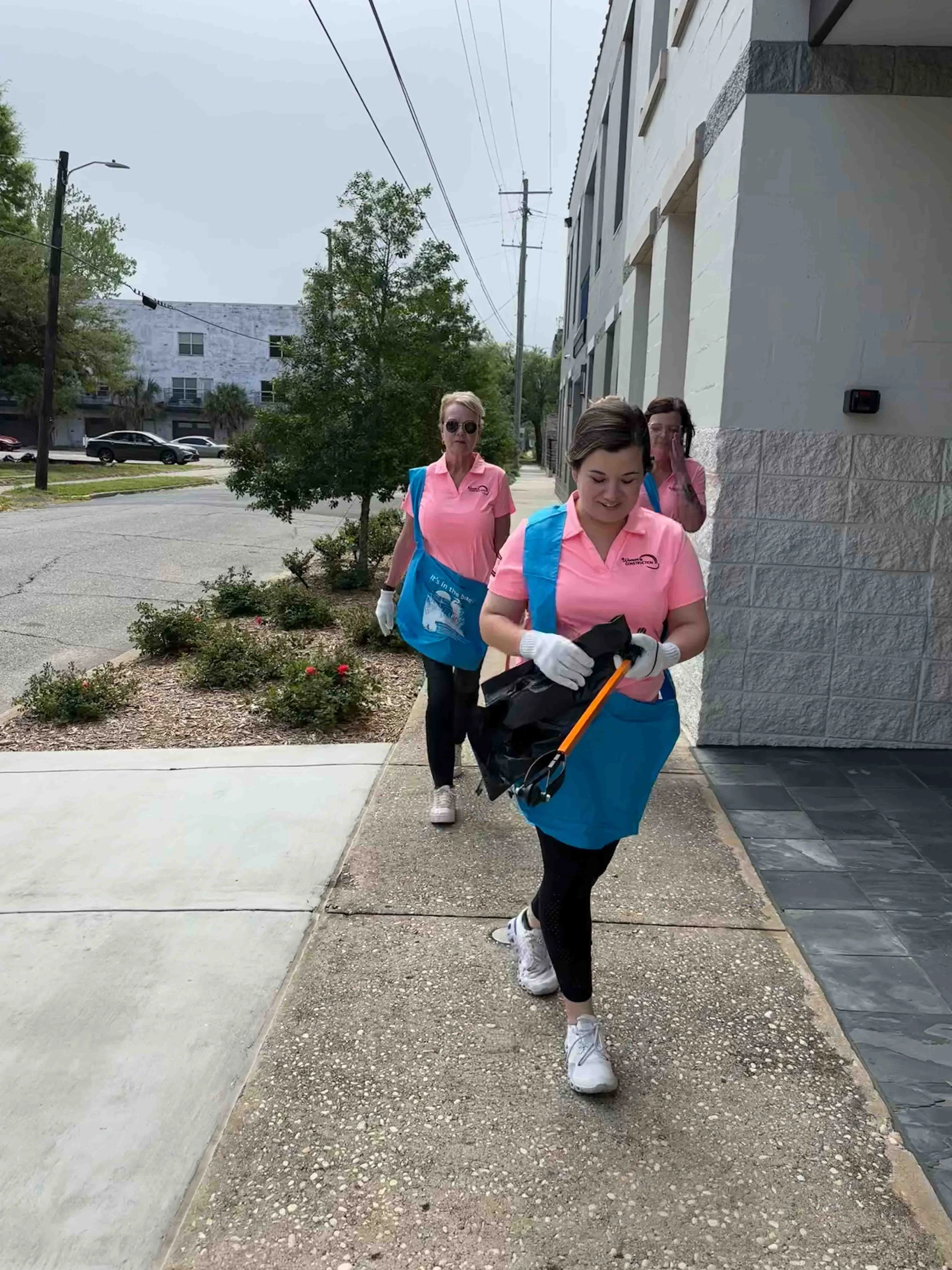 Three women dressed in pink shirts and black pants, wearing white gloves, walking together outdoors on a sidewalk near a building with greenery and parked cars in the background, some carrying blue bags.