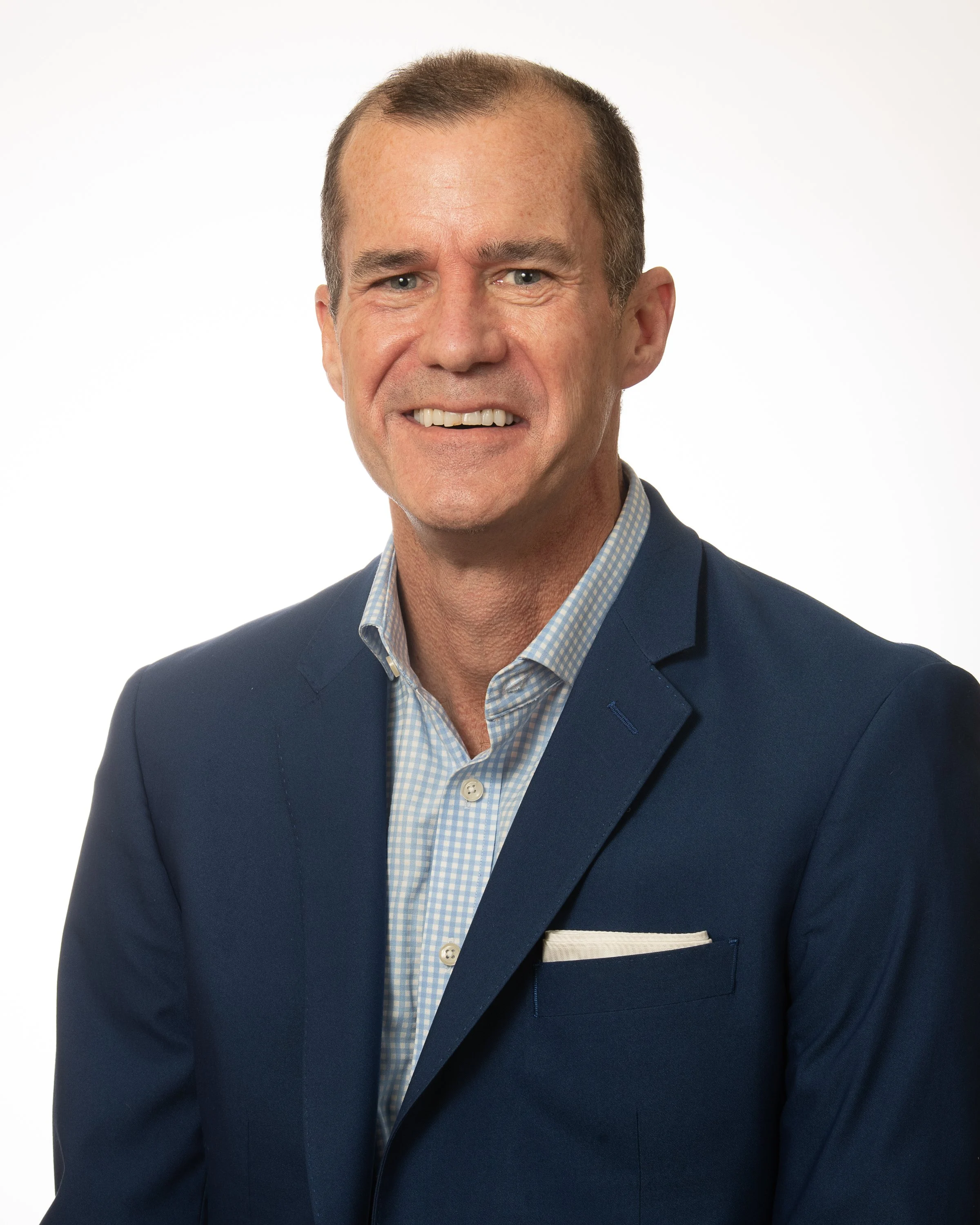 A professionally dressed man smiling, with short hair and wearing a dark suit, light blue shirt, and a tie.