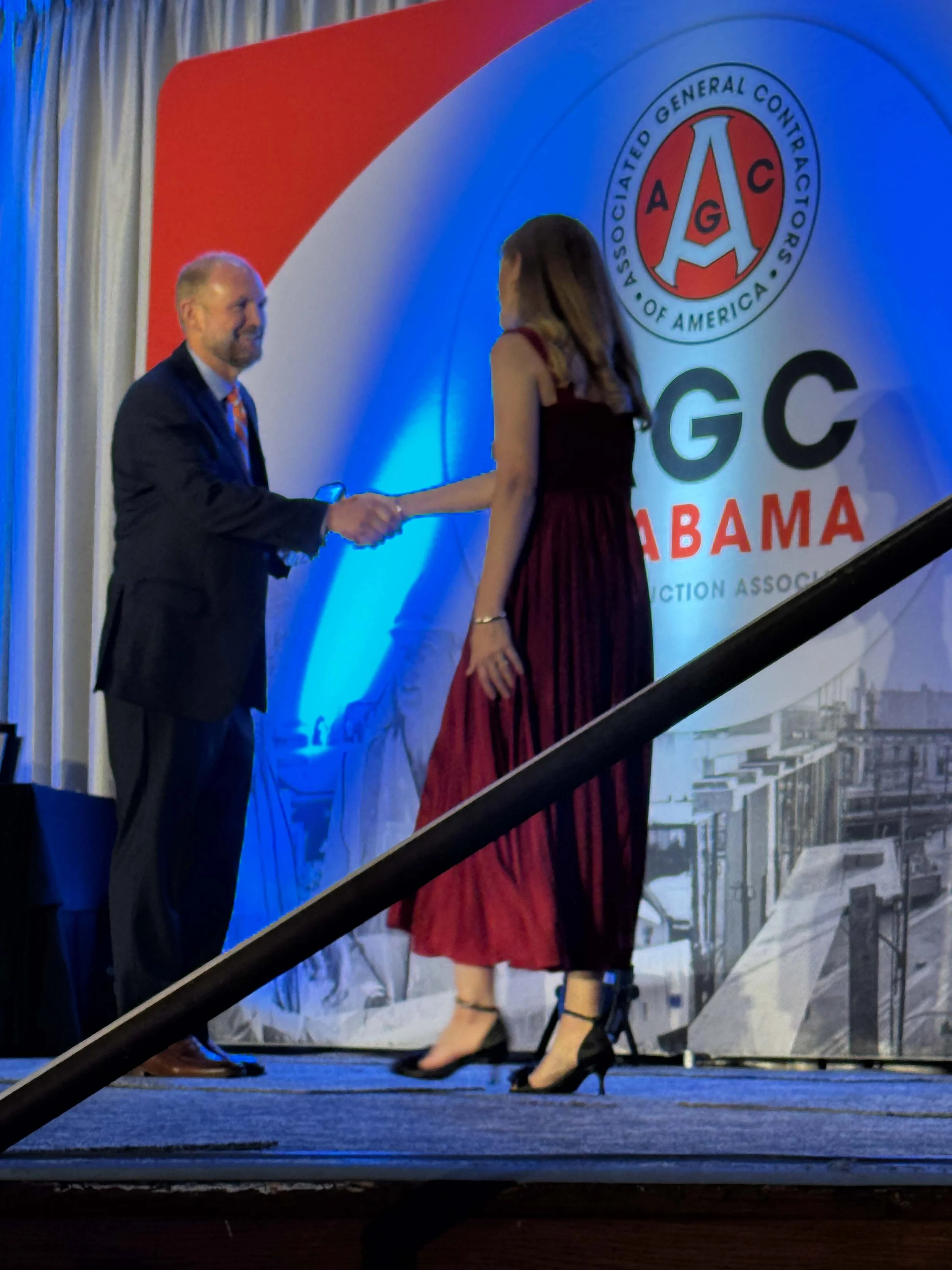 A man and a woman shaking hands on a stage at a GCA Alabama event with a large GCA Alabama logo in the background.