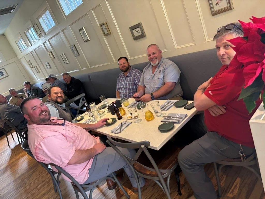 Group of six men sitting and standing around a restaurant table, smiling, with drinks and condiments, in a bright dining area with framed pictures on the wall.