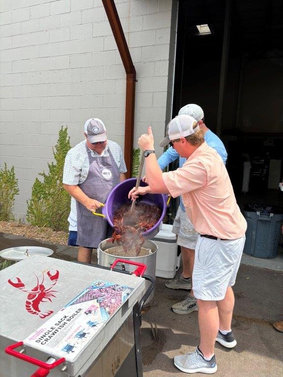 Three men preparing a seafood boil outdoors. One man is pouring the contents from a purple bucket into a pot, while another man stands behind him, and a third man is watching. They are all wearing caps and casual clothes.