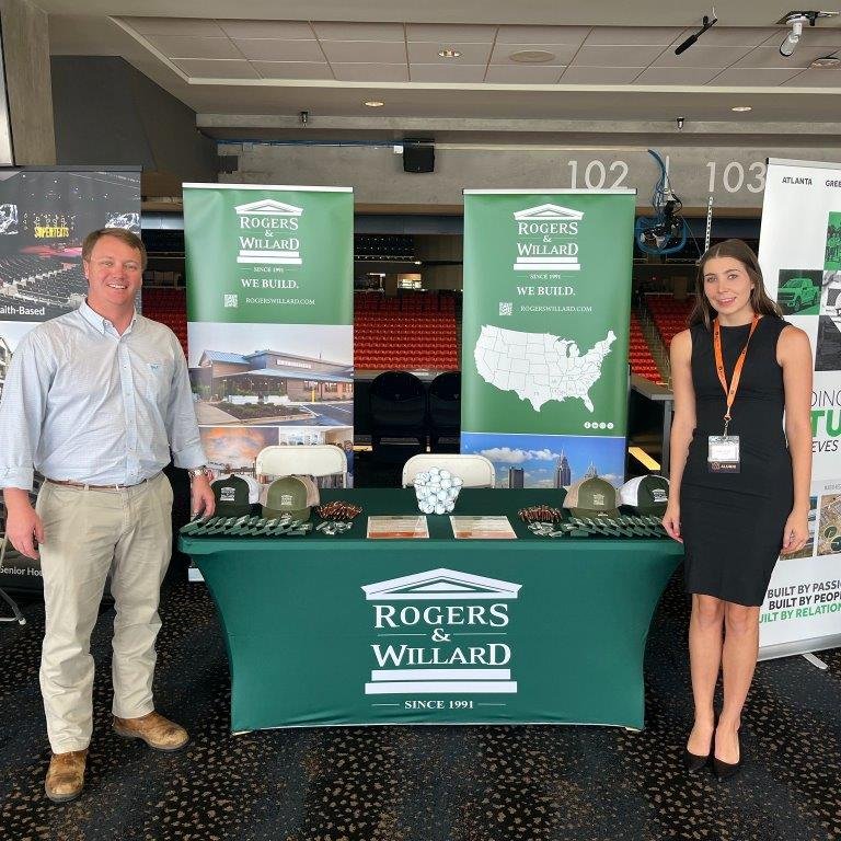 Two people standing next to a booth with Rogers & Willard banners at a trade show or conference.