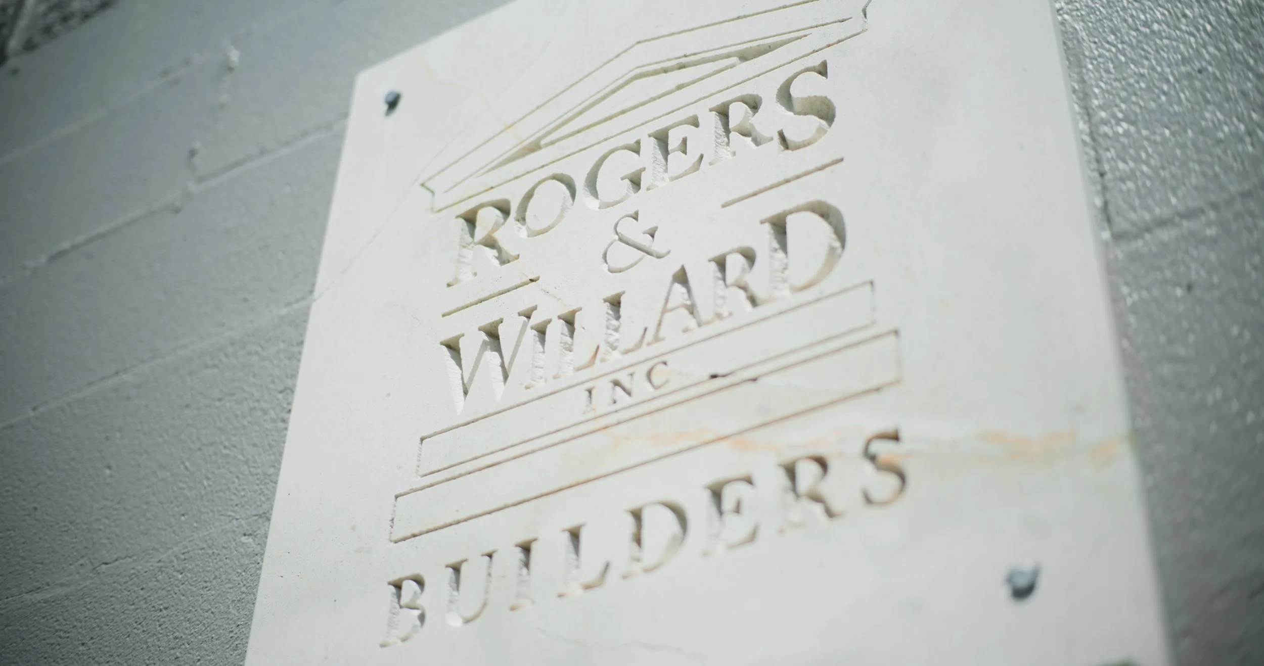 Close-up of a white carved plaque with the text "Rogers & Willard Inc" and "Builders" on a gray brick wall.