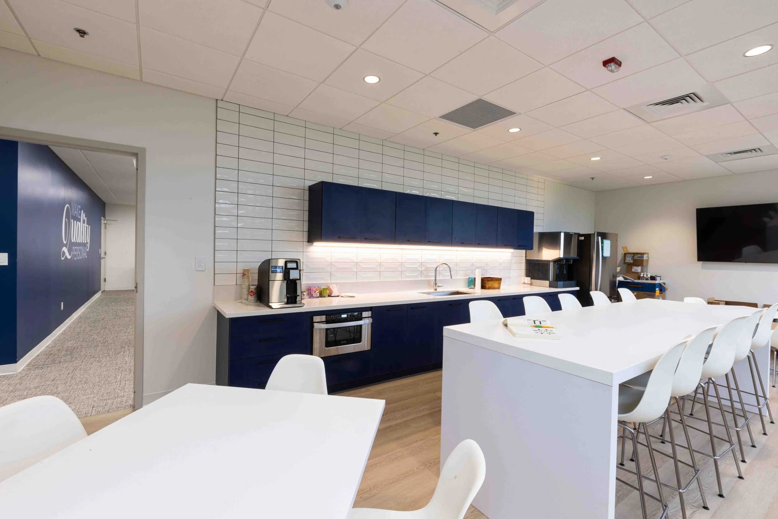 Modern office kitchen with white and navy blue cabinets, white tile backsplash, white countertop, and white chairs arranged around a white island. There is a coffee machine, a microwave, and a mini fridge. A large flat-screen TV is mounted on the wall.