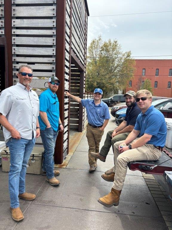 Five men standing and sitting outdoors on a sidewalk near a brick building, wearing casual and work clothes, with parked cars and trees in the background.