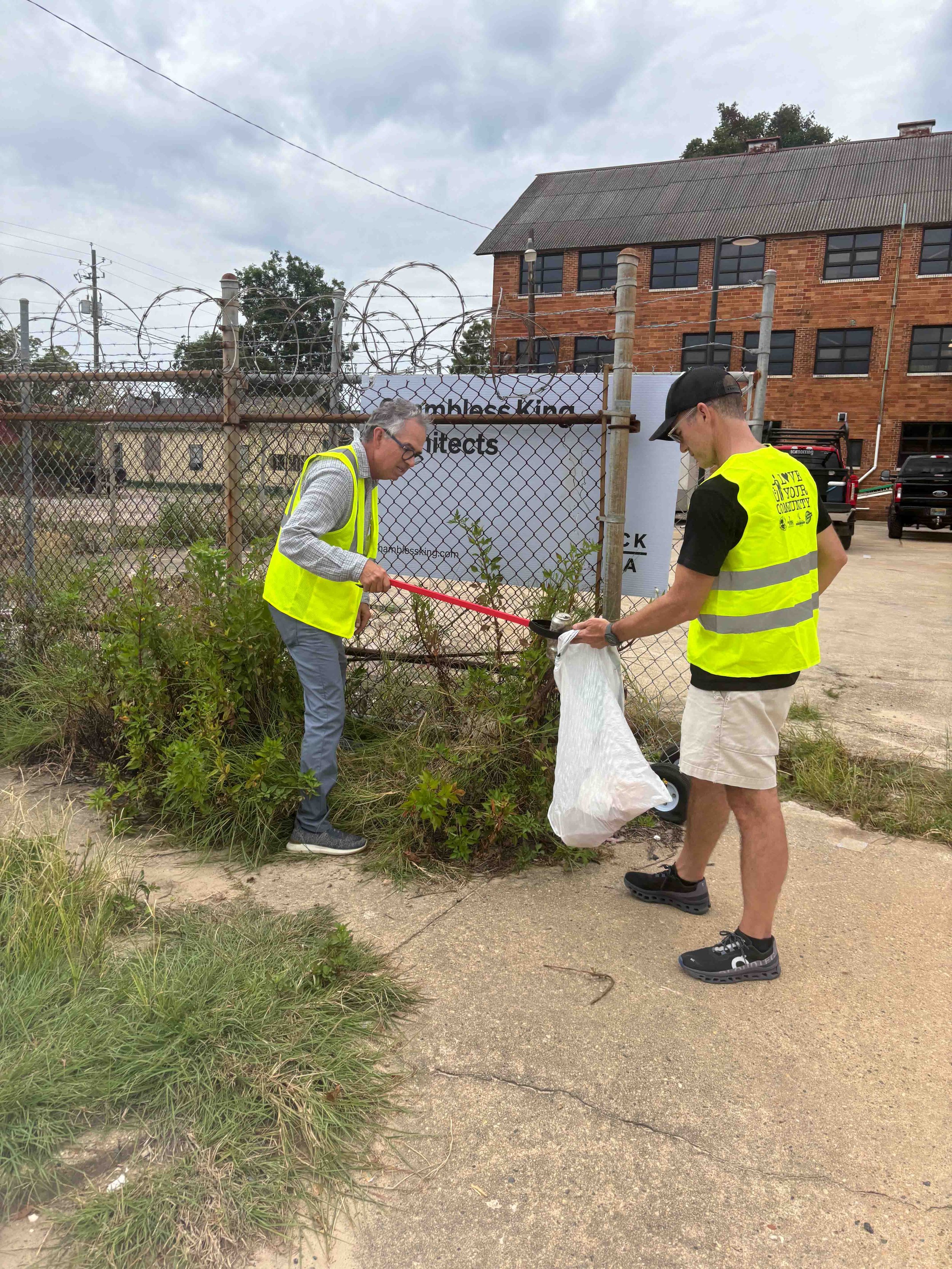 Two men wearing yellow safety vests picking up trash along a sidewalk next to a fence with a sign. One man is using a litter picker, and the other is holding a trash bag, collecting debris from the bushes.