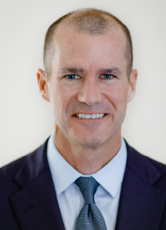 A professionally dressed man smiling, with short hair and wearing a dark suit, light blue shirt, and a tie.