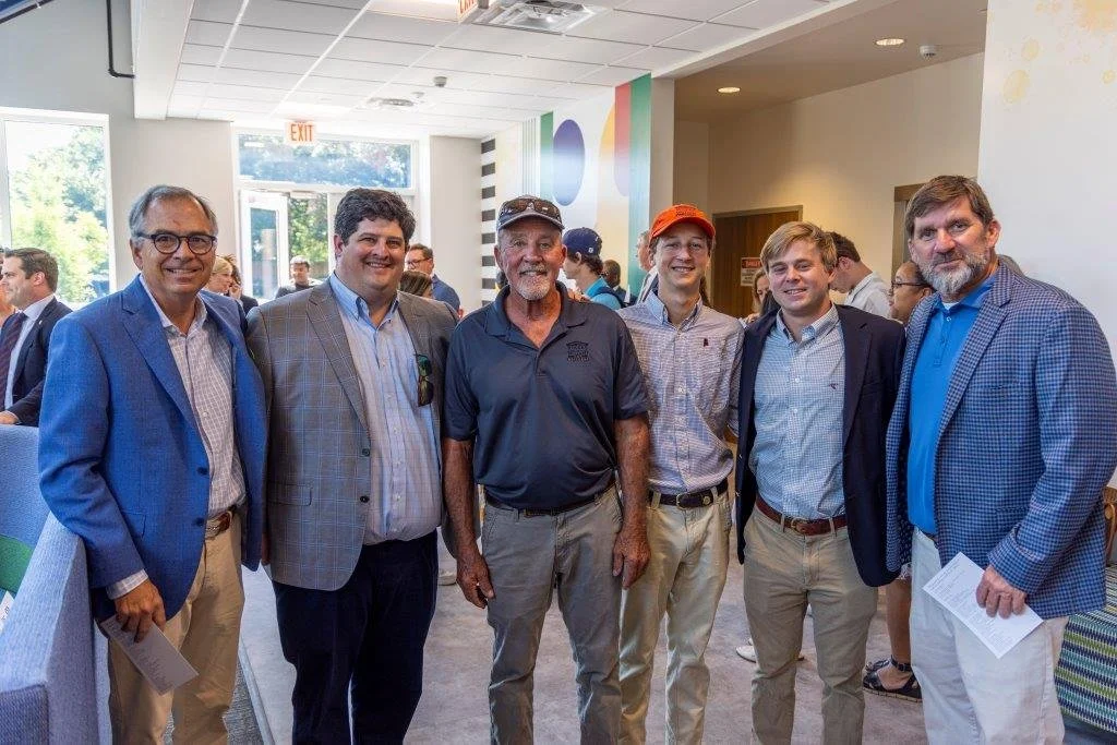 Group of six men standing indoors at an event, smiling for the camera, with other attendees in the background.