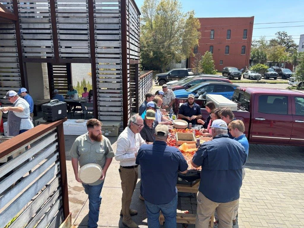 People gathered outdoors around a table with food, in a parking lot next to a building, with cars and trees in the background.