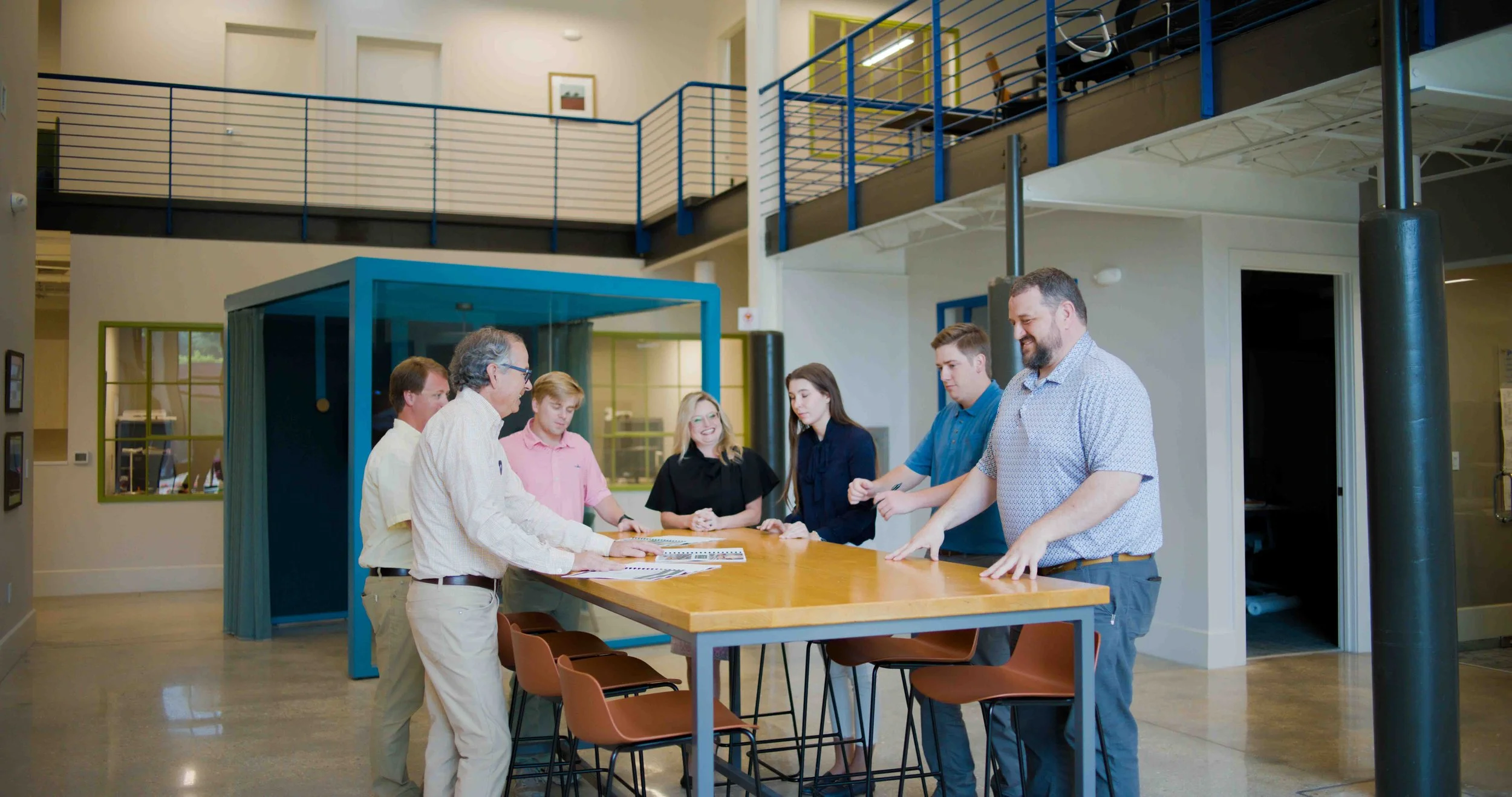 A group of eight people gathered around a wooden table in a modern office or coworking space, engaging in a discussion.