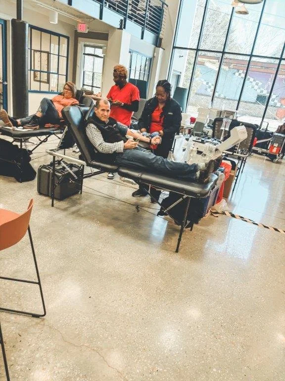 A man is lying on a medical chair donating blood or plasma, with two healthcare workers attending to him. In the background, another person is reclining on a chair, possibly waiting.